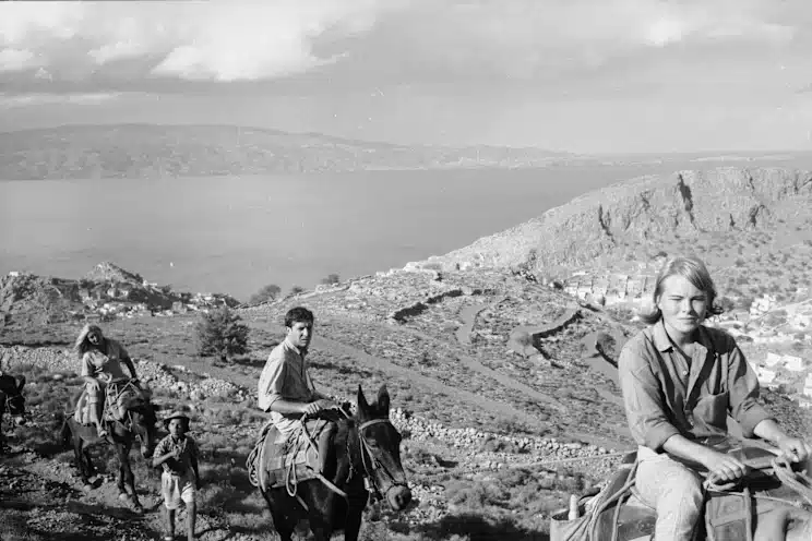 Black and white photo of Leonard Cohen and Marianne Jensen riding on donkeys over a rocky hill in Hydra.