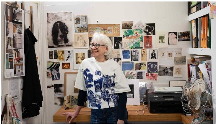 Teresa Lawrence in her studio with a large collection of her artwork on the wall behind her.
