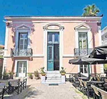 The Nafsimedon Hotel in Nafplio with pink masonry walls, tall windows and doors, and with tables and chairs and umbrellas out the front.